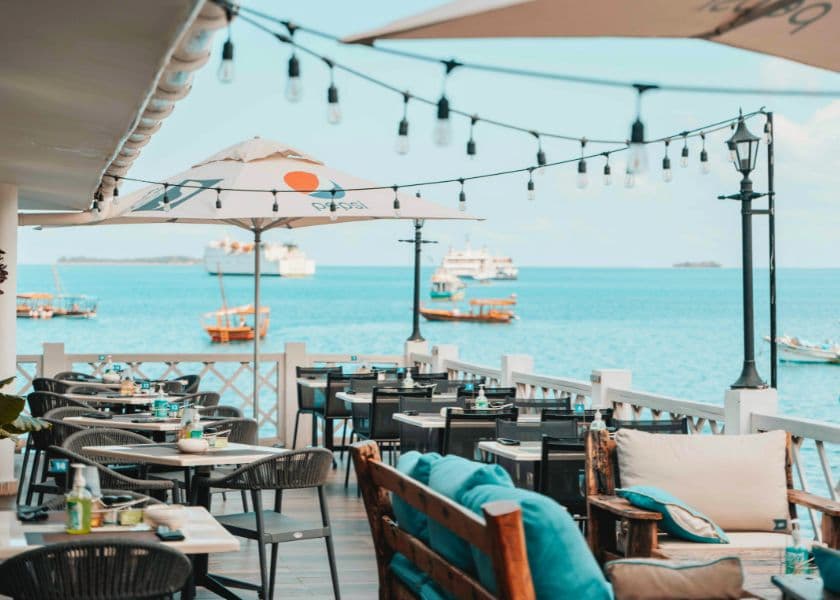 Outdoor waterfront dining area with tables, string lights, and umbrellas overlooking boats on the water. Outdoor waterfront dining area with tables, string lights, and umbrellas overlooking boats on the water.
