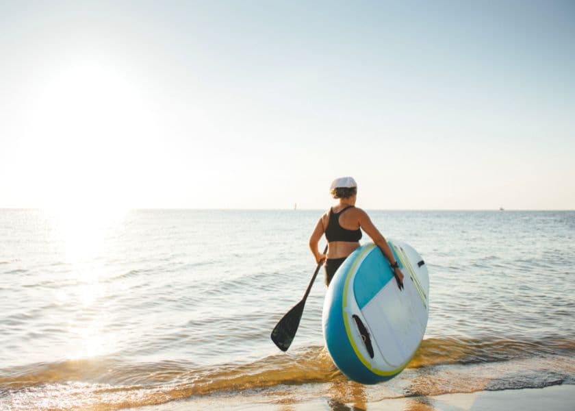 A woman walks into the water carrying a paddleboard and a paddle during a calm, sunny day at the beach. A woman walks into the water carrying a paddleboard and a paddle during a calm, sunny day at the beach.