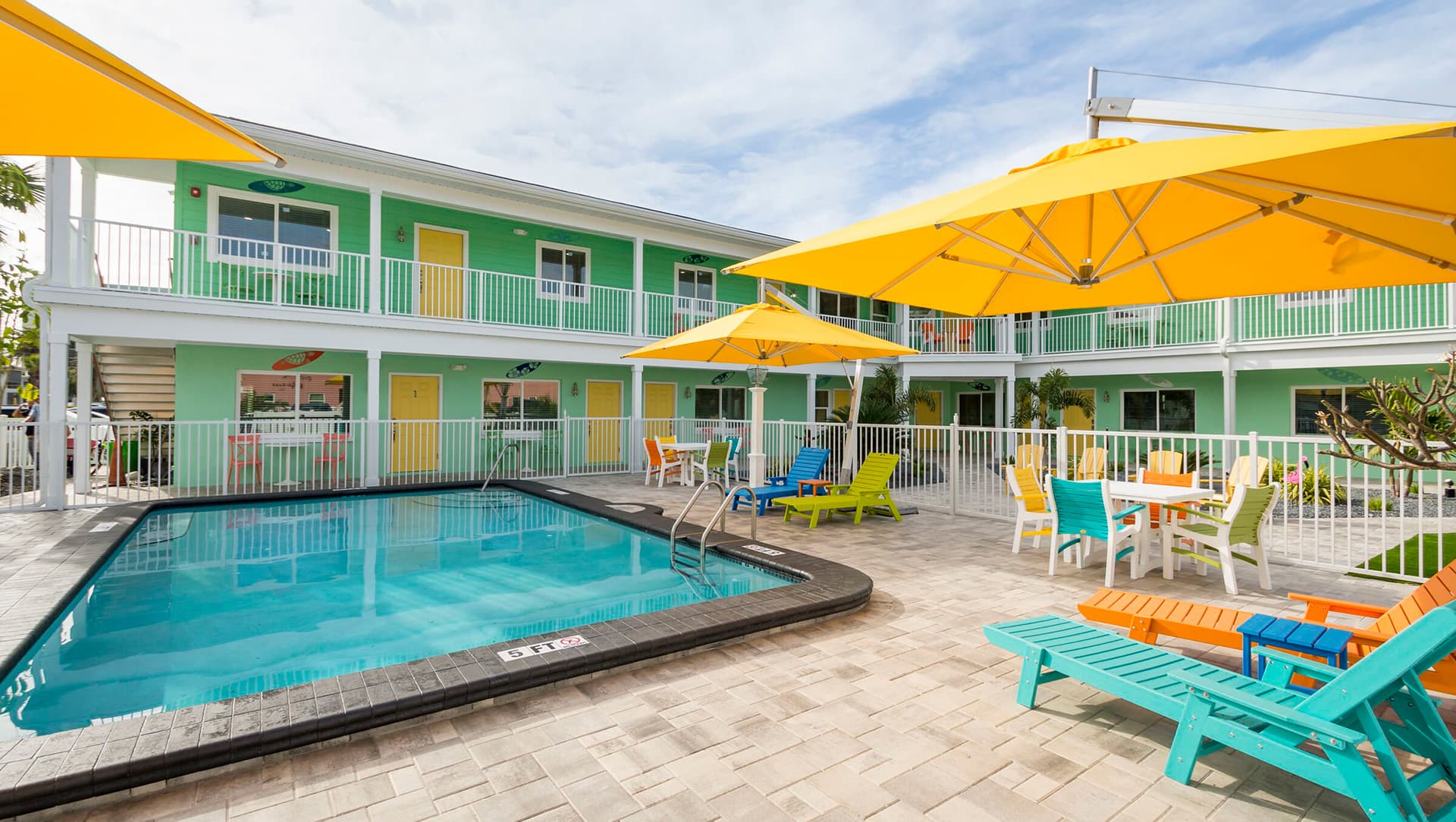 Colorful outdoor pool area with lounge chairs and umbrellas beside a green hotel.