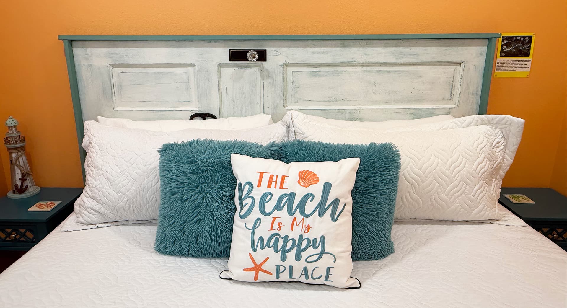 A cozy bed with white bedding and a decorative pillow that reads, "The Beach is My Happy Place," against a bright orange wall.